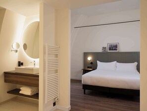 A modern hotel room with a large bed, a bedside table with a lamp, a round mirror, a built-in shelf with towels, and a white radiator. - Doubletree By Hilton Sittard (Sittard)