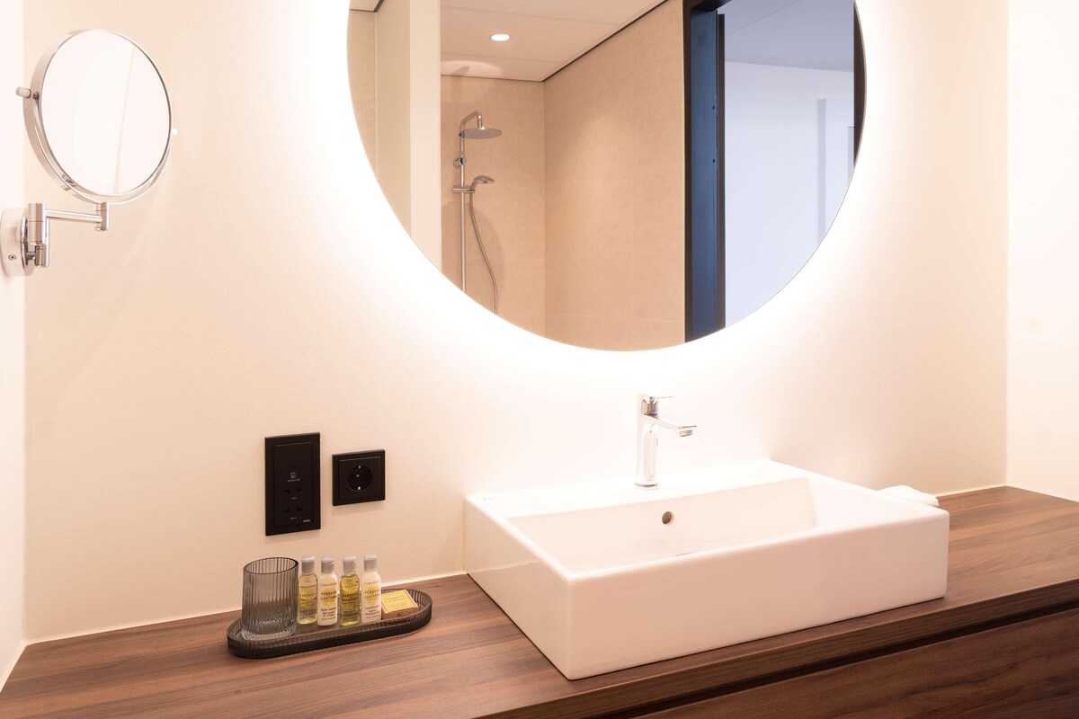 A modern bathroom with a round mirror, a rectangular sink, and a shower visible in the reflection.