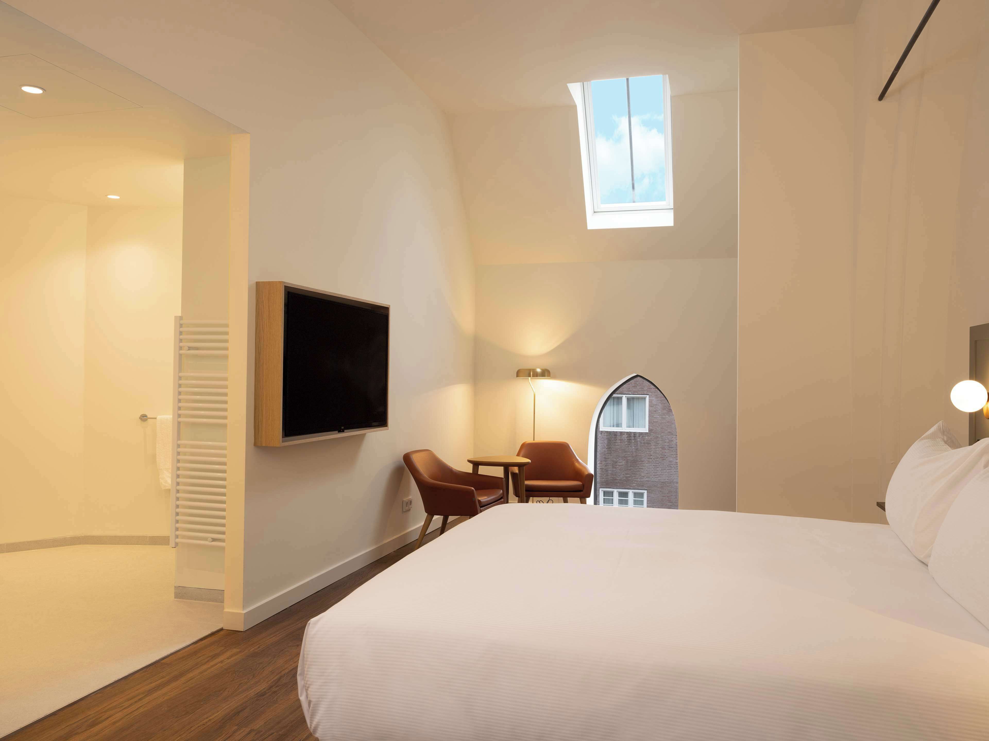 Ett hotellrum med en säng, en tv som är monterad på väggen, ett litet bord med stolar, en lampa och en unik arkitektonisk detalj som liknar ett hus.