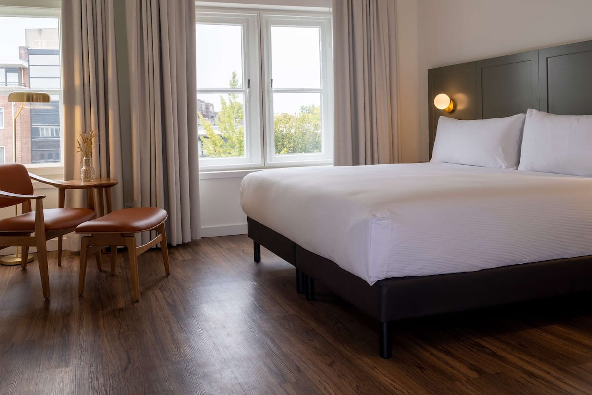 A hotel room with a large bed, a desk with two chairs, a small table, and a window with curtains.