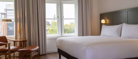 A hotel room with a large bed, a desk with two chairs, a small table, and a window with curtains.