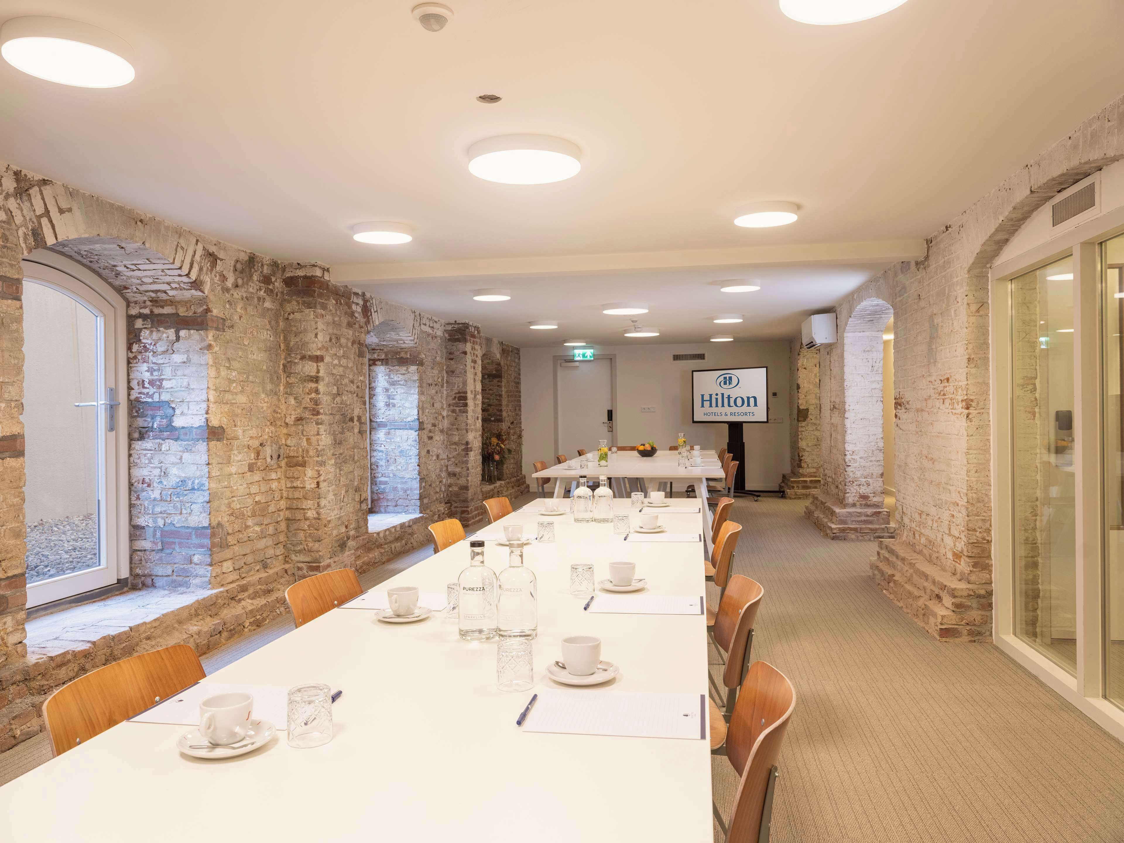 A conference room with a long table set for a meeting, brick walls, and a Hilton logo on the wall.