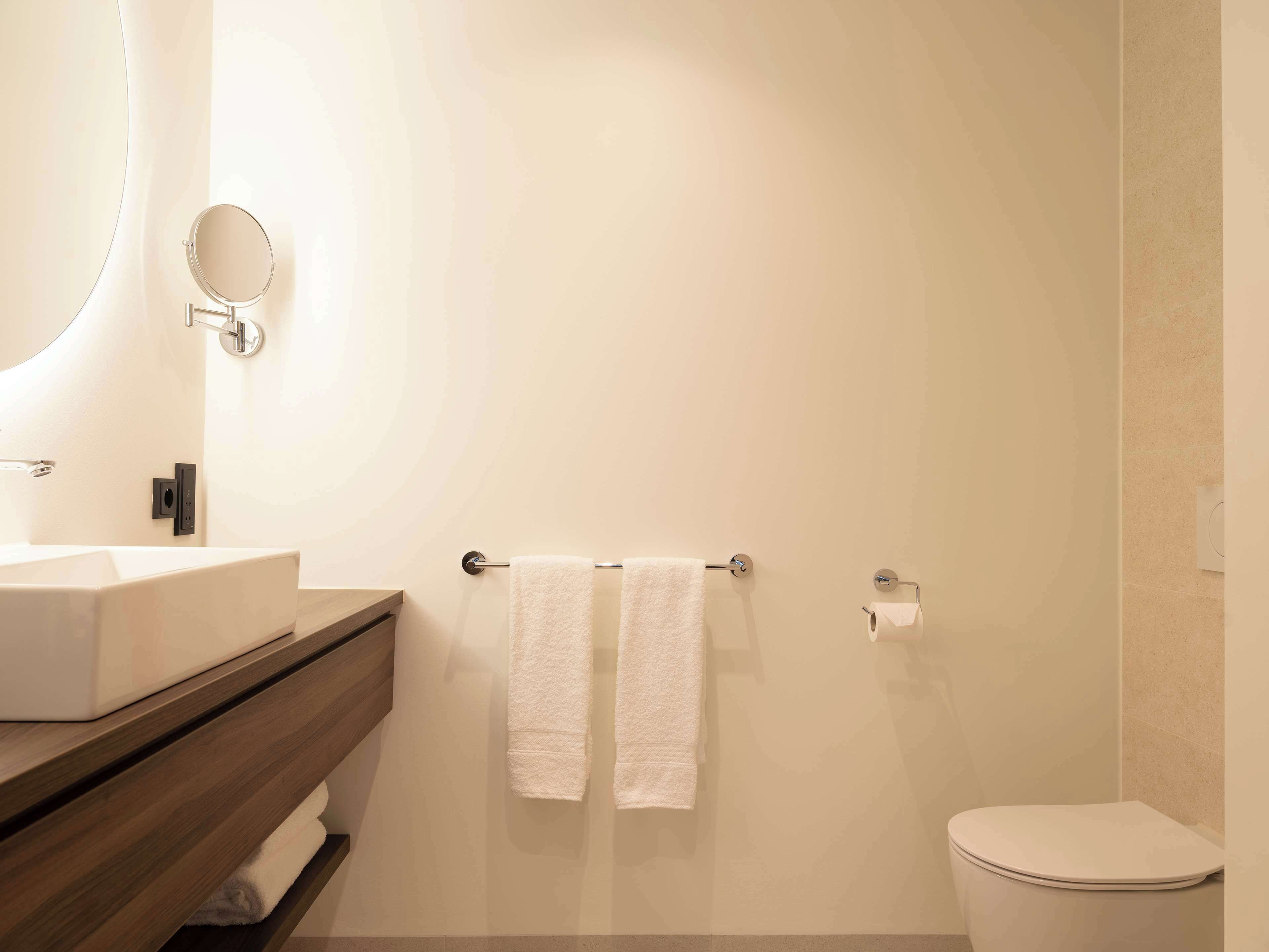 A modern bathroom with a rectangular sink, a round mirror, two white towels on a towel rack, and a toilet.