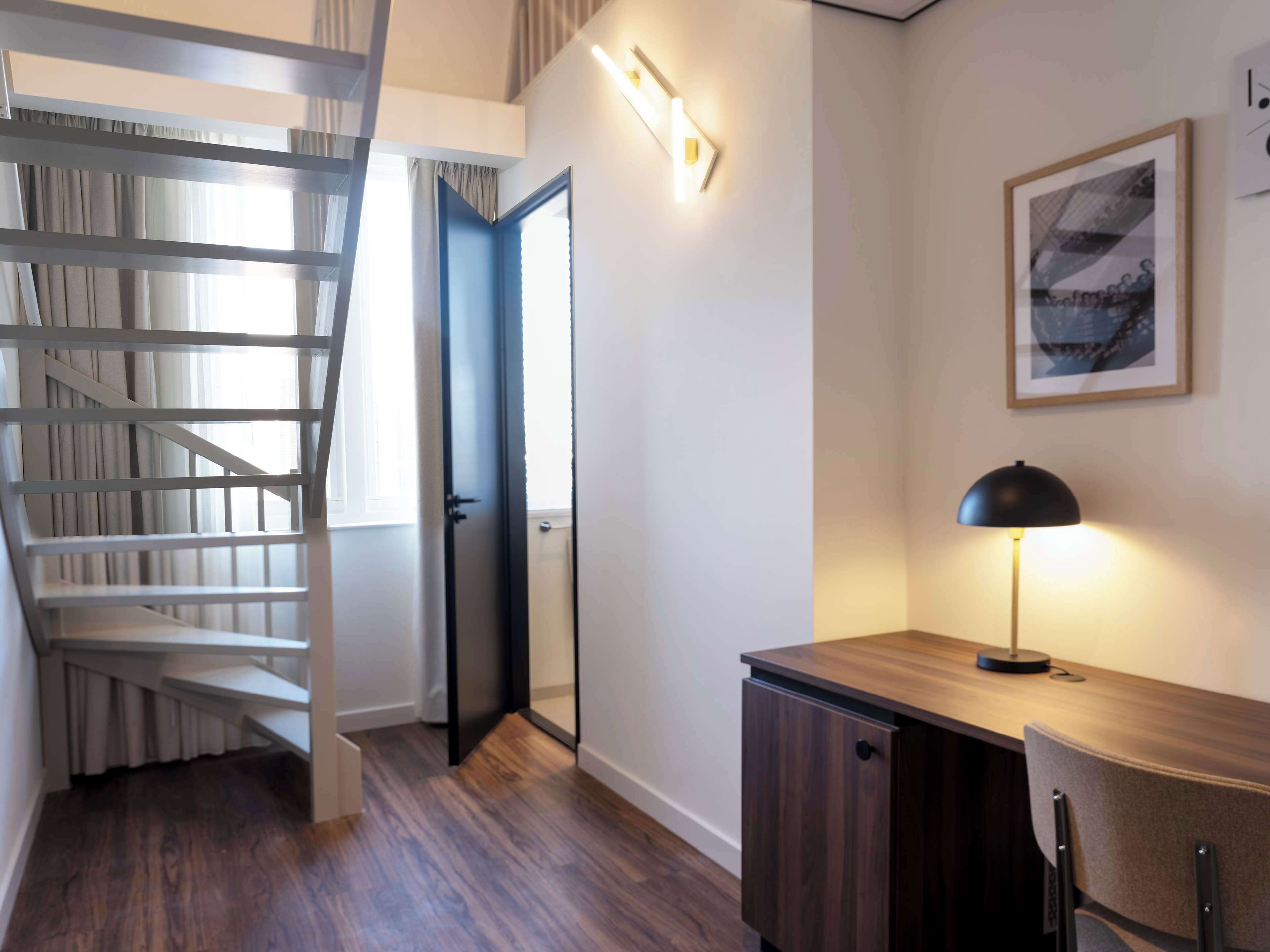 Een moderne kamer met een houten bureau, een lamp, een lijst met een afbeelding en een trap naar een ander deel.