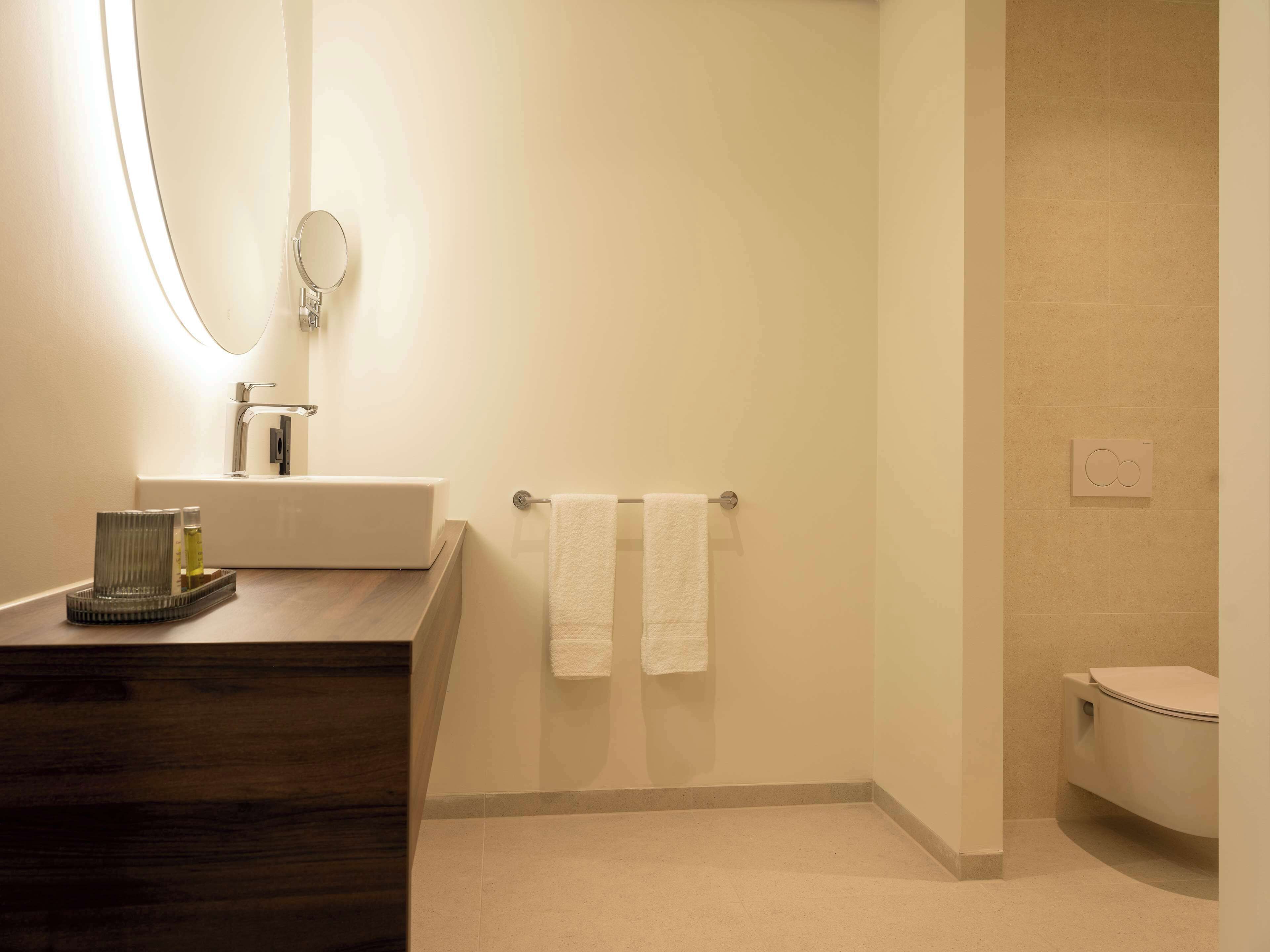 A bathroom with a wooden vanity, a mirror, a towel rack with two towels, a toilet, and a wall-mounted light fixture.