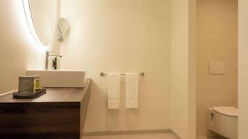 A bathroom with a wooden vanity, a mirror, a towel rack with two towels, a toilet, and a wall-mounted light fixture.