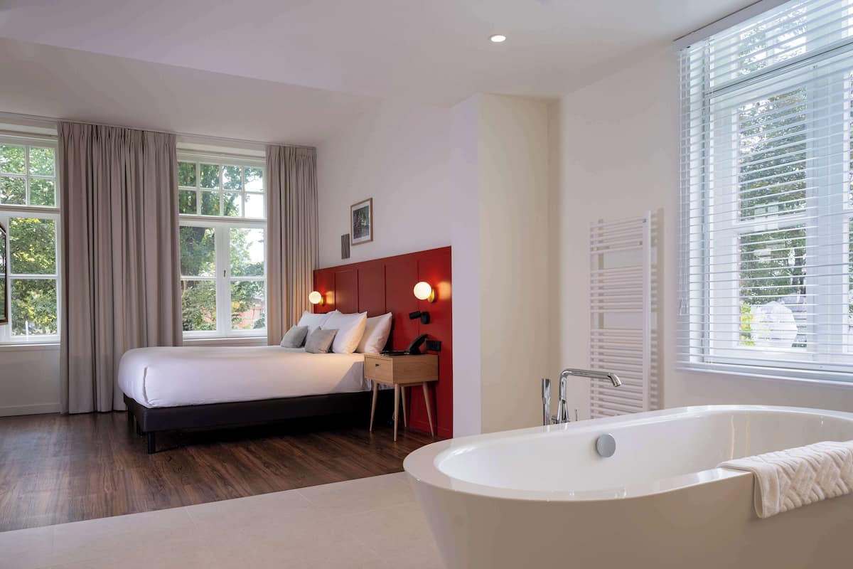 A modern bedroom with a large bed, a bedside table, a red headboard, a wall-mounted light, and a window with curtains.