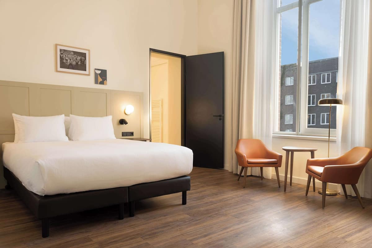 A hotel room with a large bed, two chairs, a small table, a nightstand, and a window with curtains.