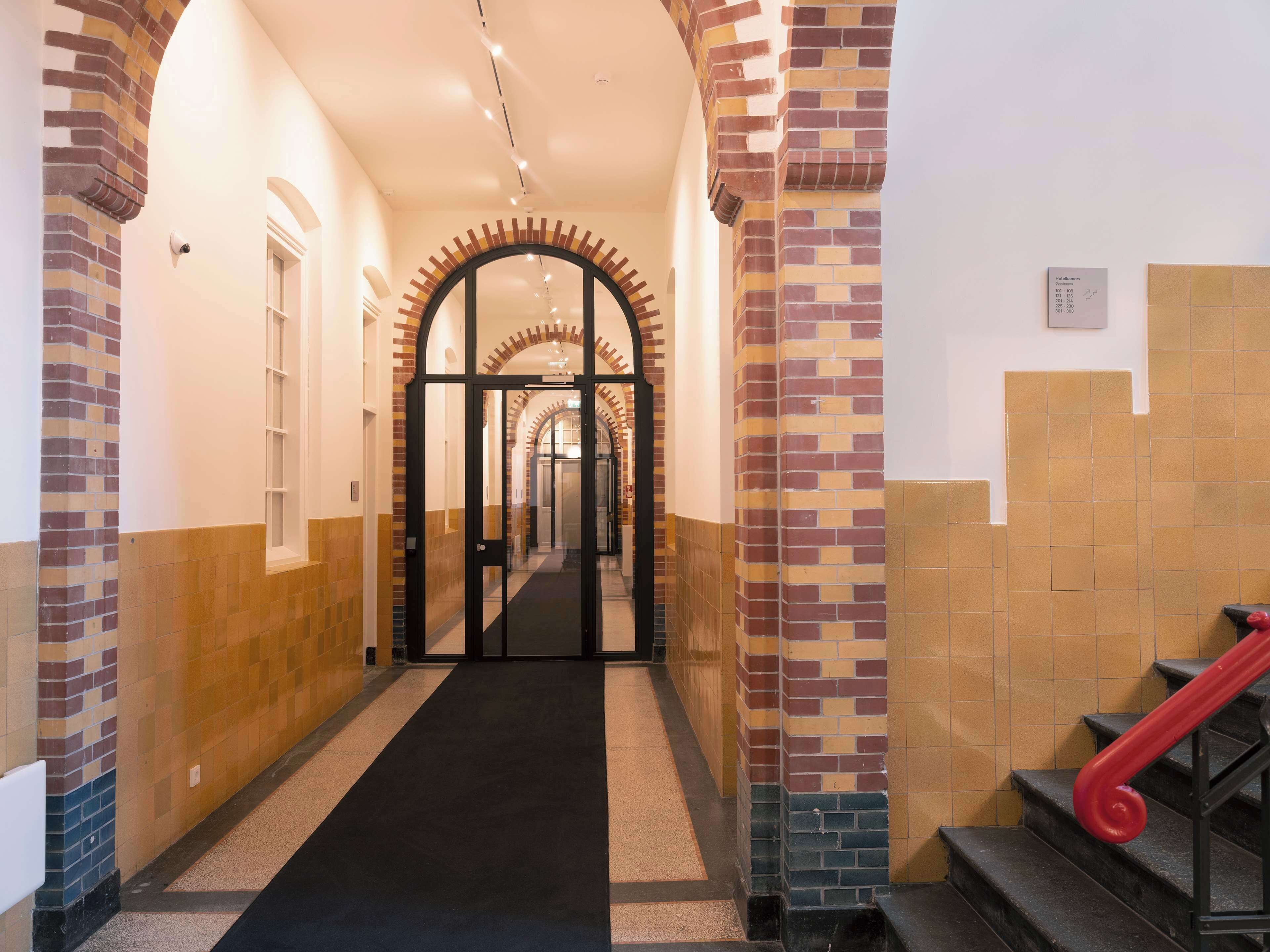 Un pasillo de hotel con alfombra negra, paredes de ladrillo y azulejos beige, y una escalera roja.