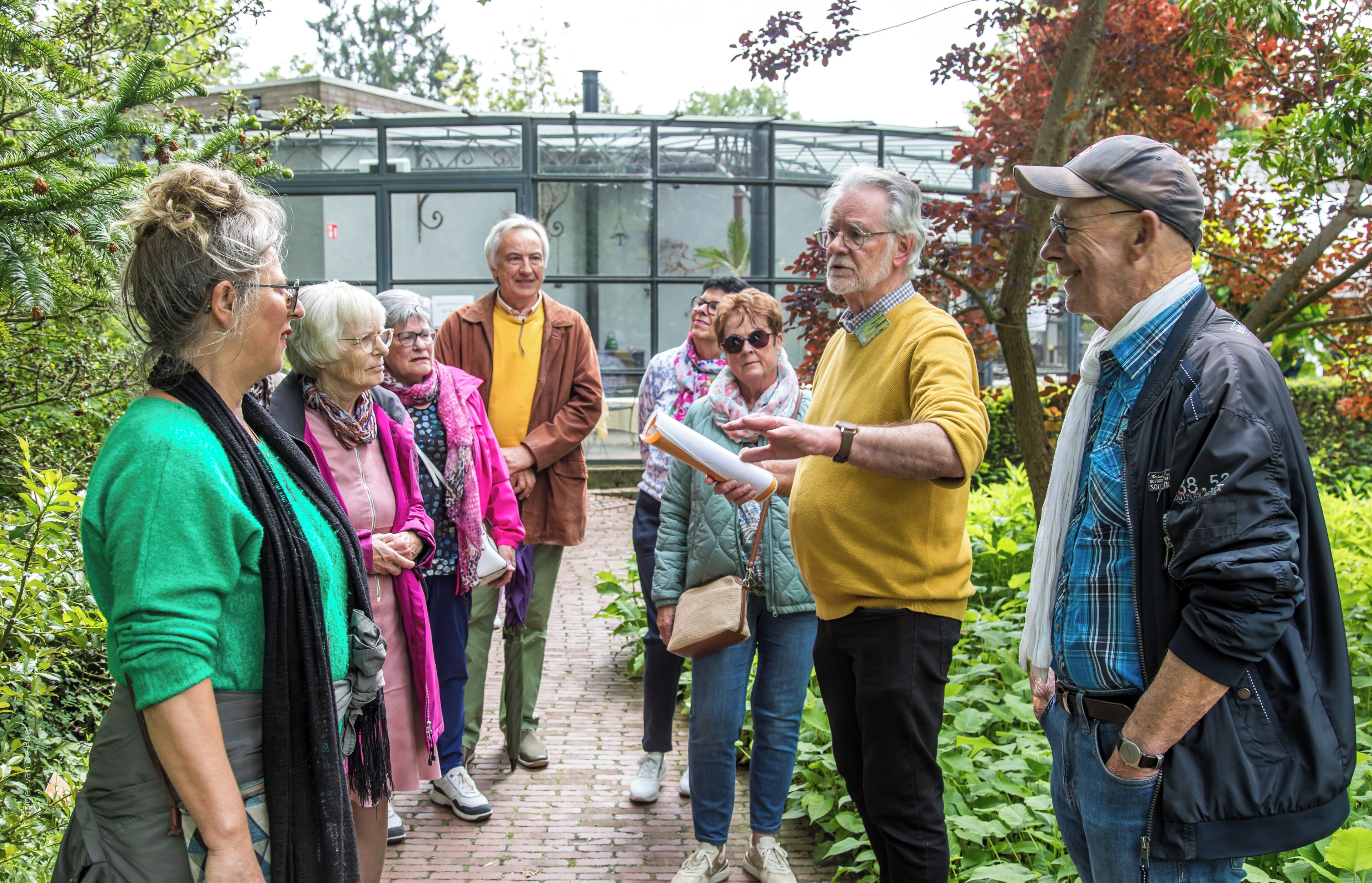 A group of people standing on a brick pathway in a garden with a glass greenhouse structure in the background.