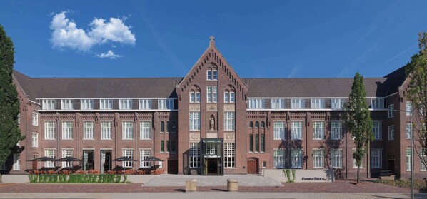 A large brick building with multiple windows, a central entrance, and outdoor seating with umbrellas. - Doubletree By Hilton Sittard (Sittard)