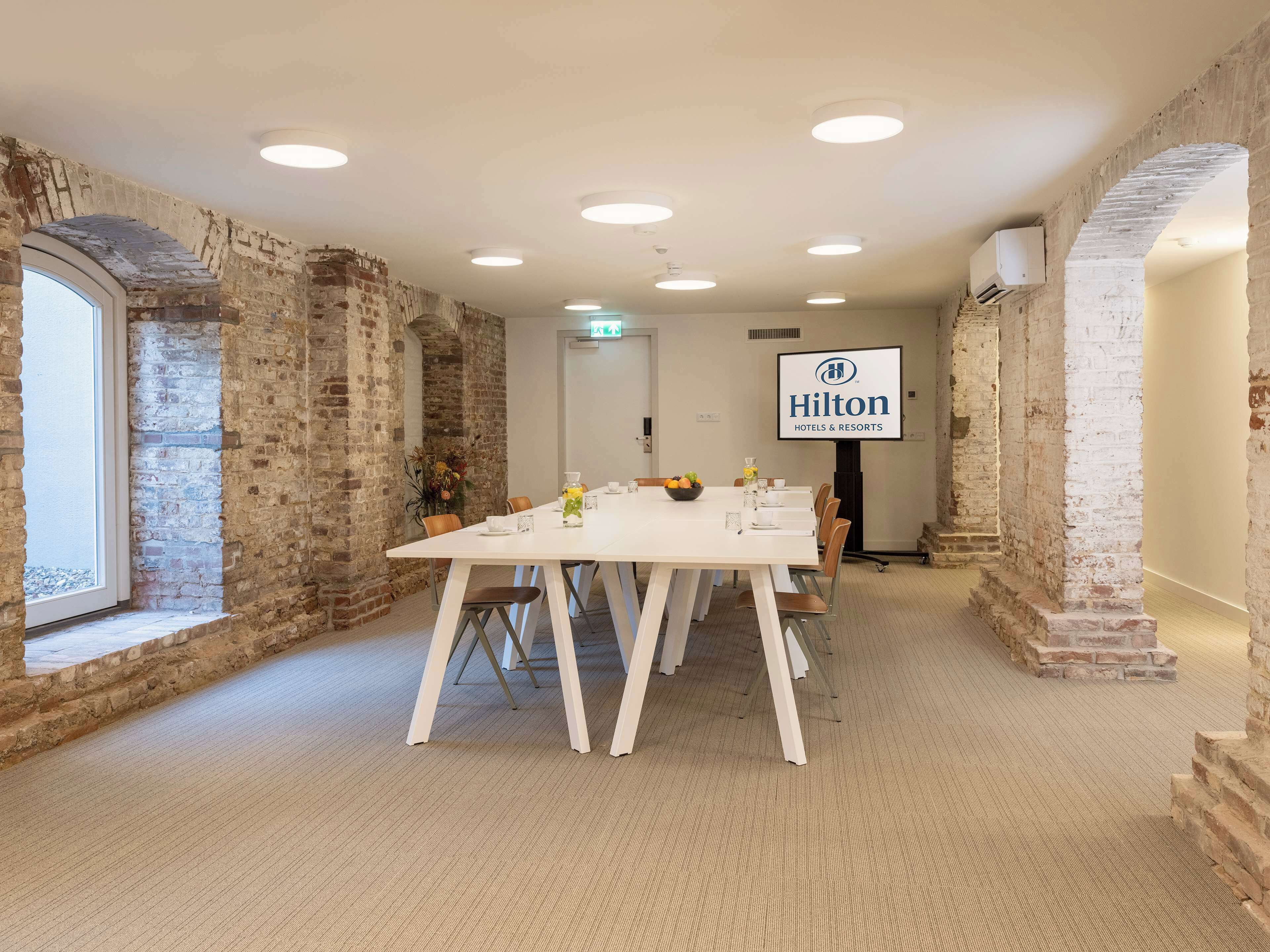 A conference room with a long table, white chairs, and a Hilton logo on the wall.