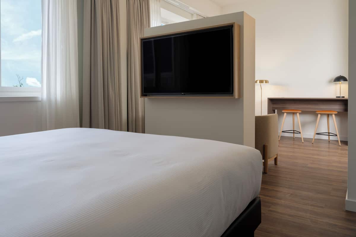 A hotel room with a large bed, a TV mounted on the wall, a desk with two stools, and a window with curtains.