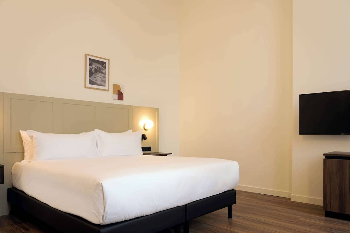 A neatly made bed with white linens, a bedside table with a lamp, a framed picture on the wall, and a TV mounted on the wall.