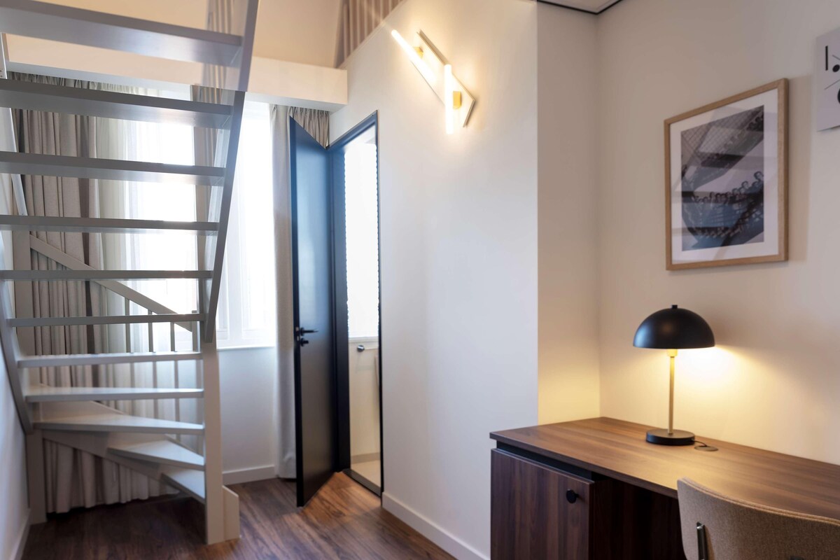 Una escalera con barandilla blanca, una puerta que da a otra habitación, un escritorio con lámpara y un cuadro en la pared.