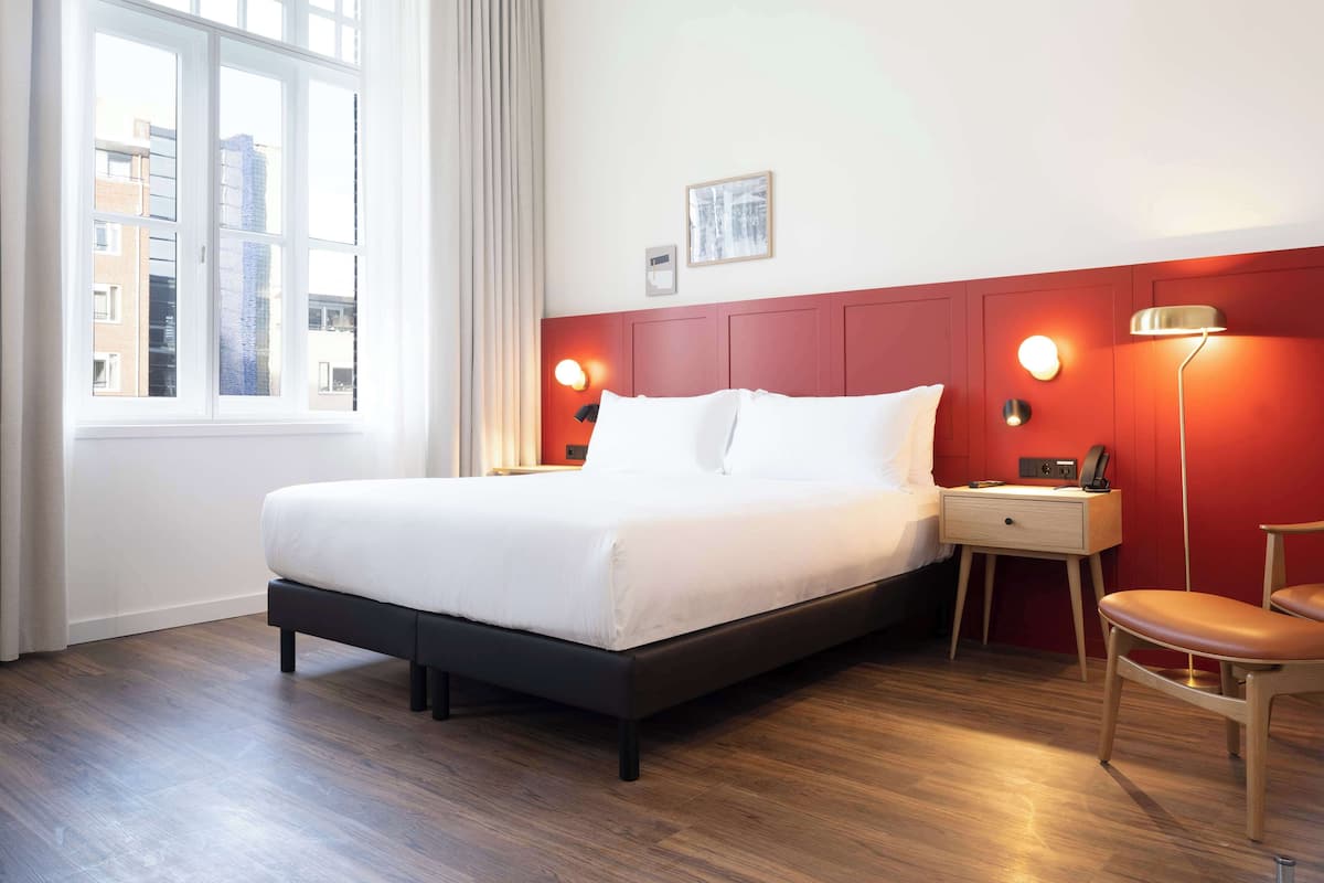 A hotel room with a bed, a nightstand, a chair, a window with curtains, and a red accent wall.