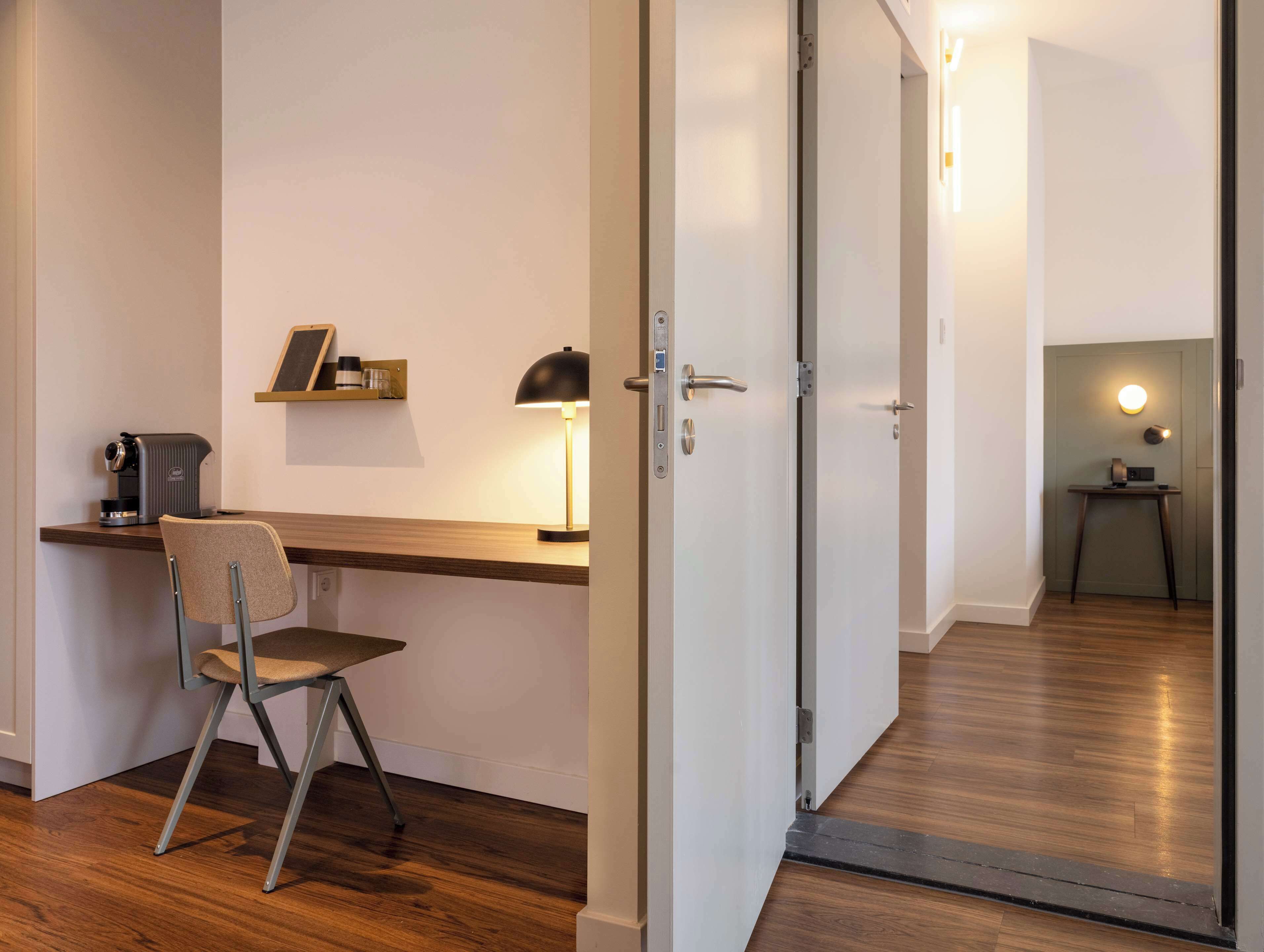 Una habitación moderna con un escritorio de madera, una silla, una lámpara de escritorio y una puerta que da a otra habitación.