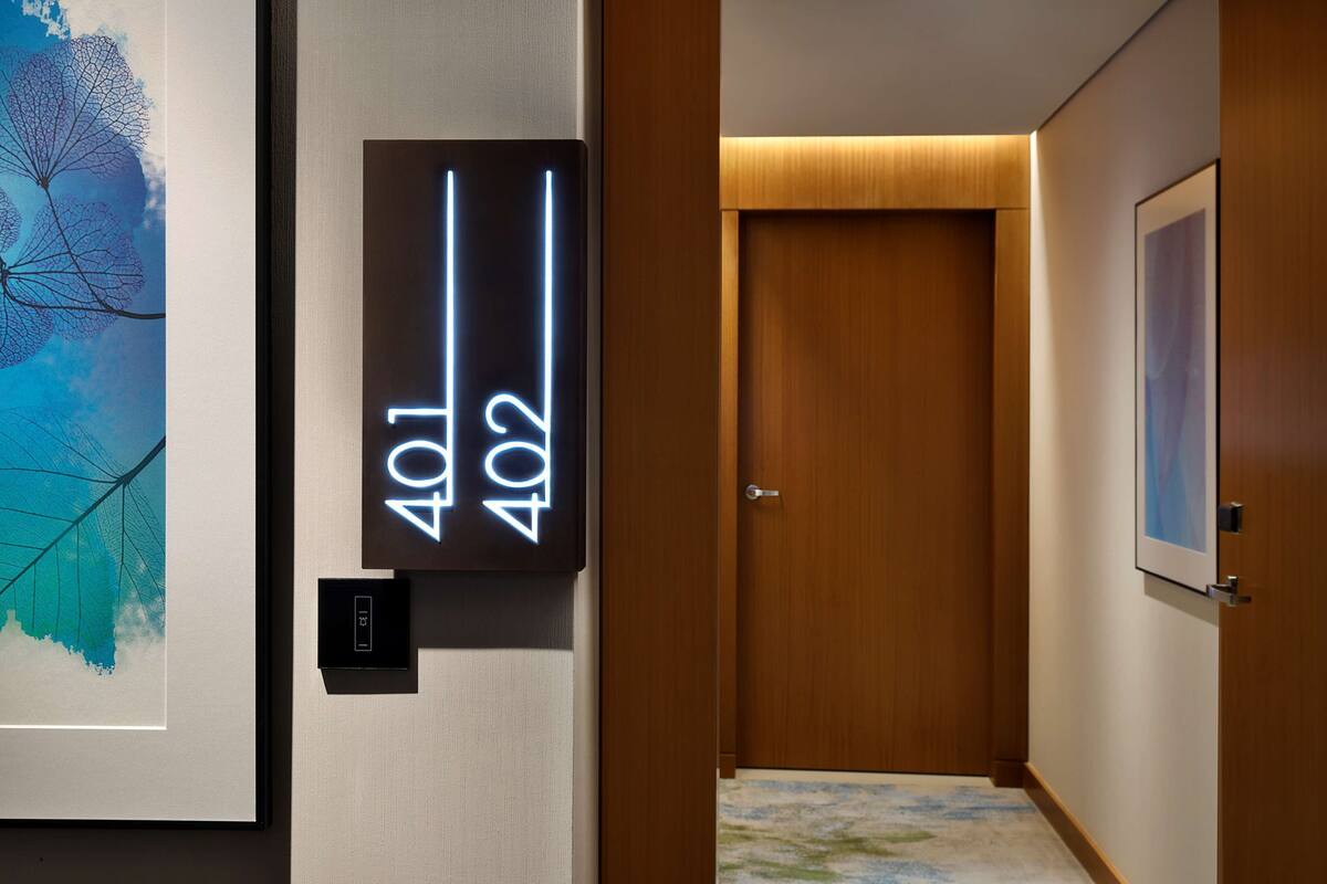 Een hotelhal met een digitaal scherm waarop kamer nummers 401 en 402 staan, een ingelijst kunstwerk aan de muur en een houten deur aan het einde.