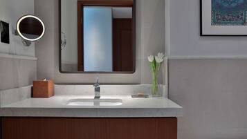 Une salle de bain avec un comptoir en marbre, un miroir rond avec un éclairage intégré, un meuble en bois en dessous et une œuvre d’art encadrée accrochée au mur.