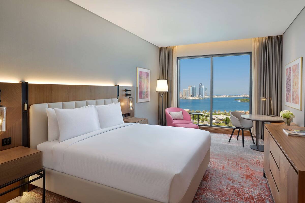 Een hotelkamer met een groot bed, een bureau, een stoel en uitzicht op de stad en zee.
