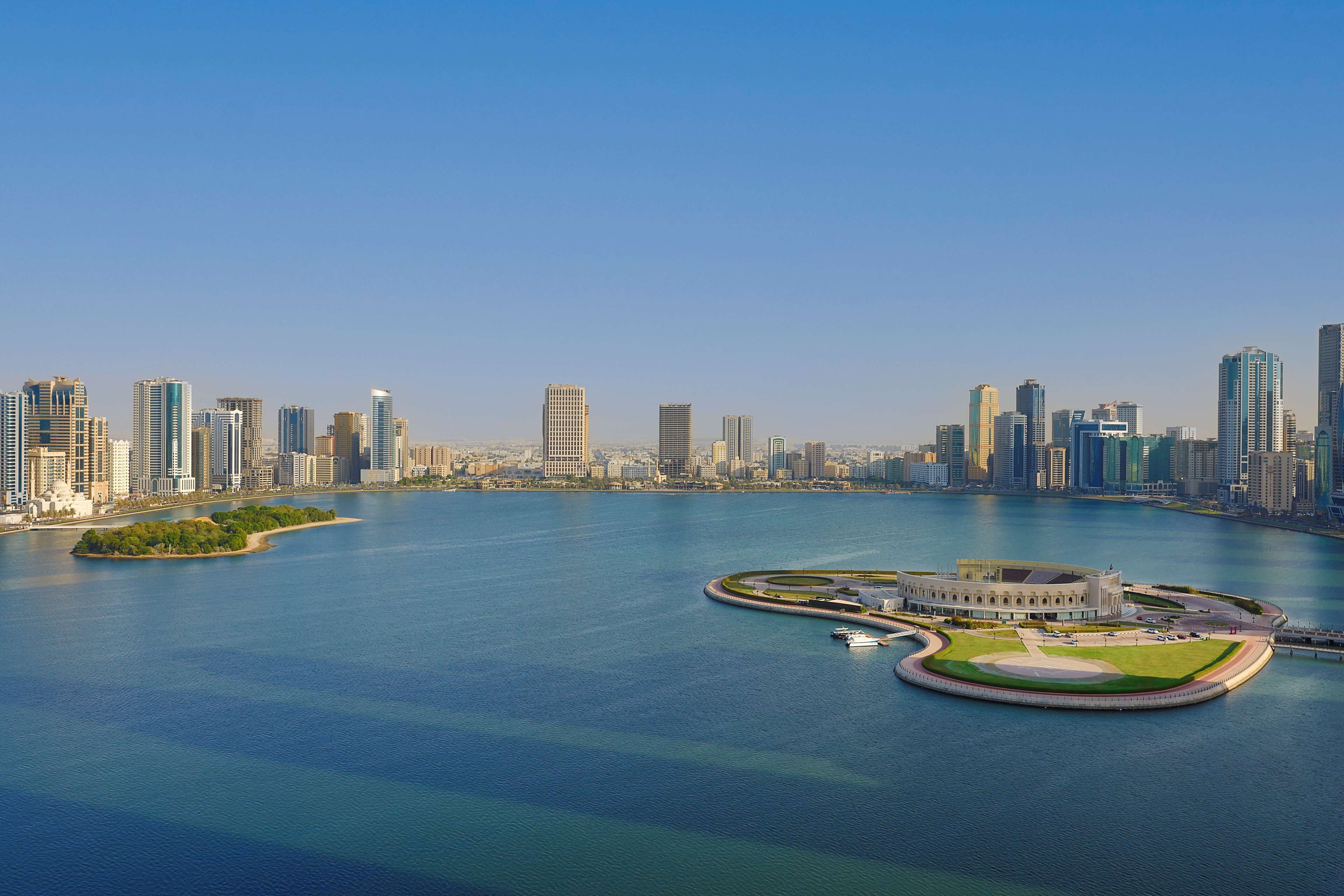 Un horizonte urbano frente al agua con edificios modernos altos, un puerto deportivo y una isla circular distintiva con un edificio en el centro.