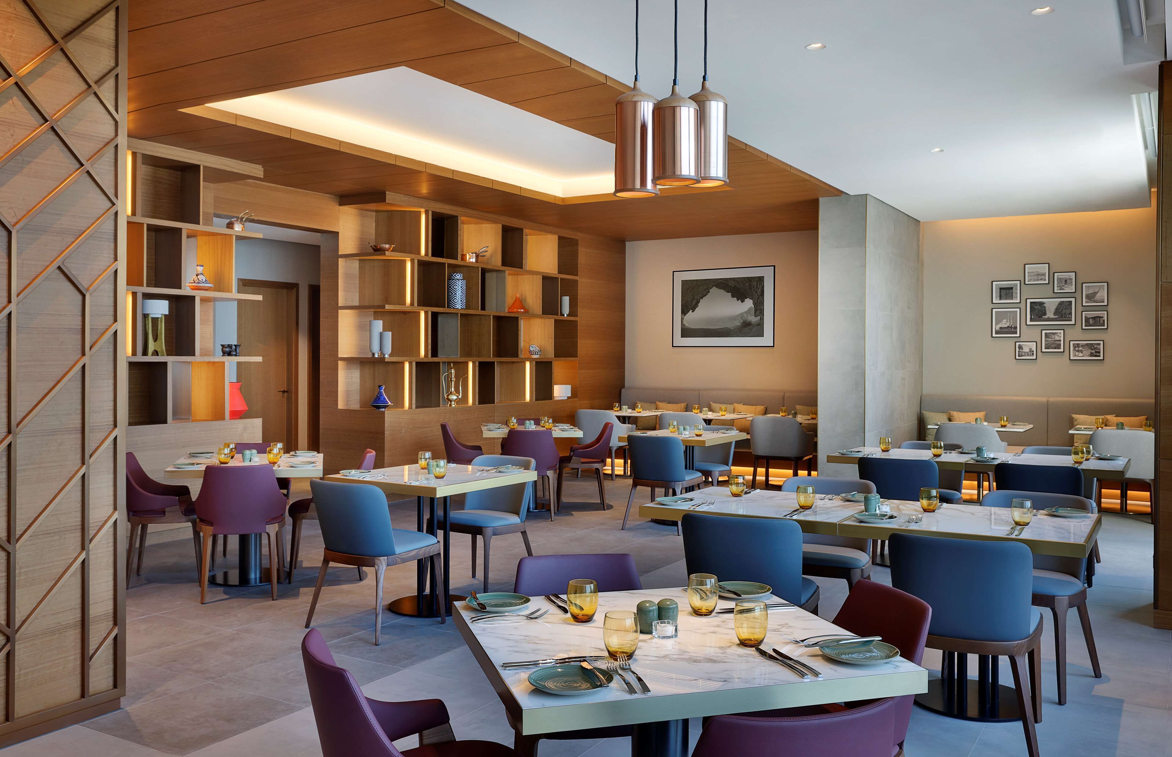 A modern restaurant with multiple dining tables and chairs, wooden shelves, and a ceiling with recessed lighting.
