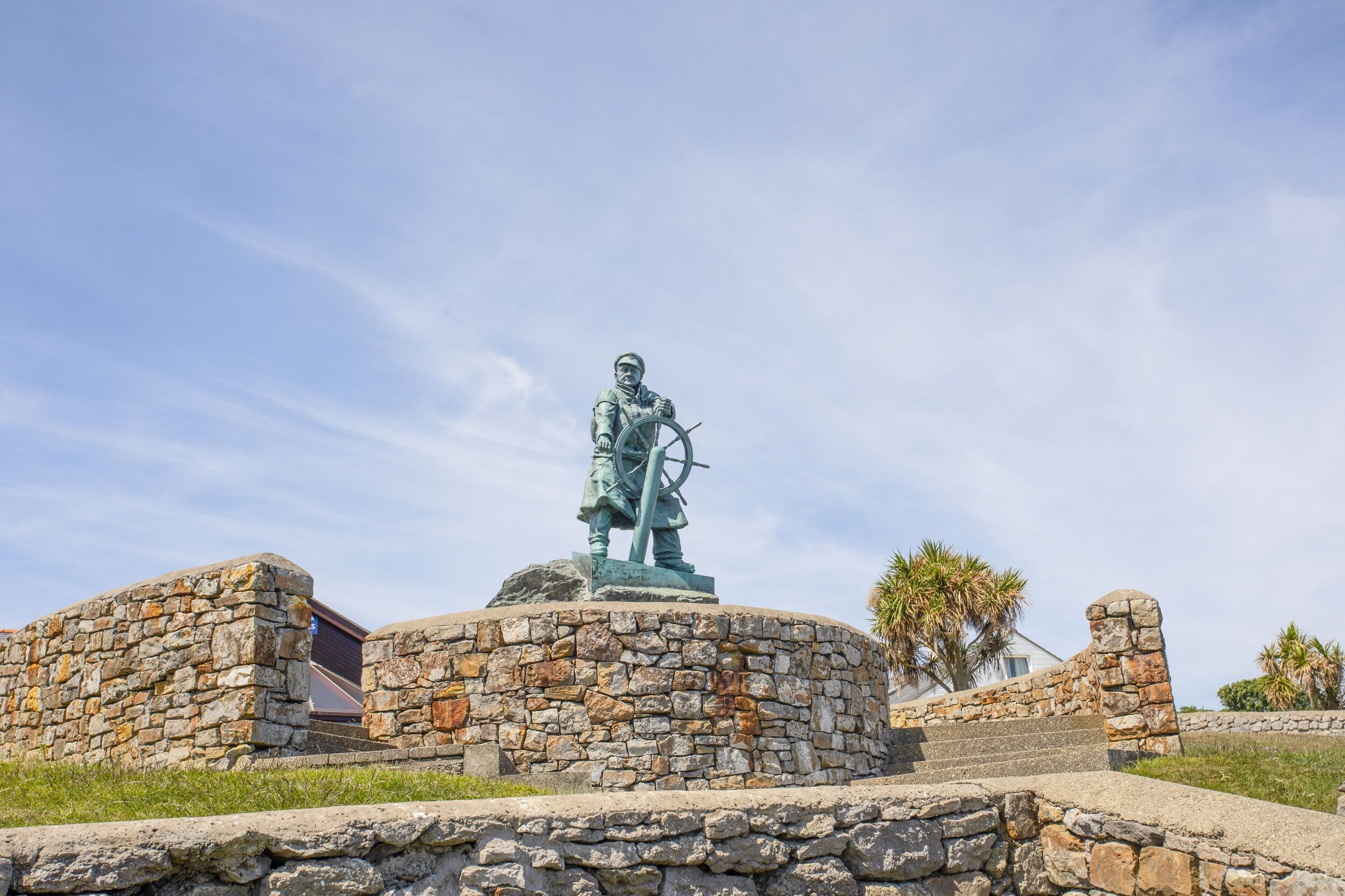 Een standbeeld van een historische figuur op een stenen sokkel, omringd door stenen muren en palmbomen.