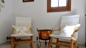 Deux fauteuils à bascule avec des coussins blancs et des motifs floraux brodés, un pot en cuivre posé sur un support, et une fenêtre avec un cadre en bois.