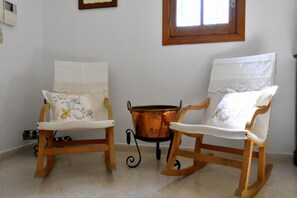 Dos mecedoras con cojines blancos y bordado floral, una olla de cobre sobre un soporte y una ventana con marco de madera.