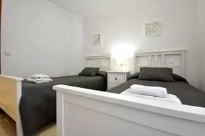 Una habitación de hotel con dos camas individuales, una mesita de noche con lámpara y otra mesita más pequeña con un reloj.