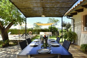Un área de comedor al aire libre con una mesa dispuesta para seis personas, rodeada de sillas, bajo una pérgola sombreada.