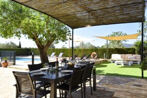 An outdoor patio with a dining table set for six, surrounded by chairs. There is a pool in the background, and a covered seating area with a sofa and chairs.