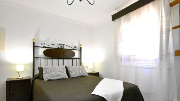 A neatly made bed with a headboard labeled "R", two bedside lamps, and a window with sheer curtains. - 44262 Finca Els Xiprers (Porreres)