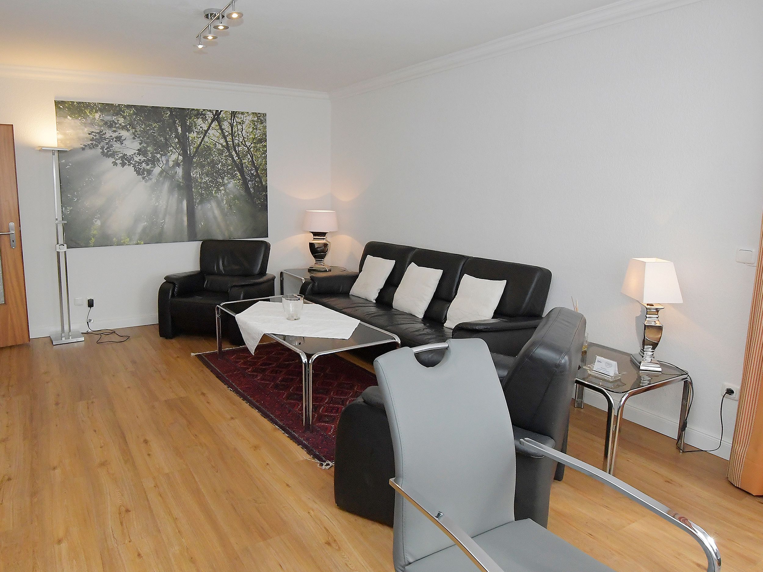 A modern living room with a black leather sofa, a grey armchair, a glass coffee table, and a red rug. There is a painting of trees on the wall and a lamp on a side table.