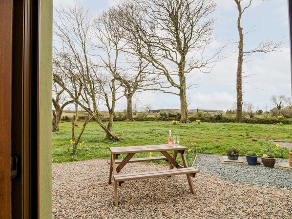 A wooden picnic table with a bottle and glasses on a gravel area with potted plants.
