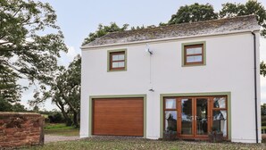 A two-story building with a white exterior and green trim, featuring a large garage door and a set of double doors with glass panels. - Bramblewood Cottage (Wigton)