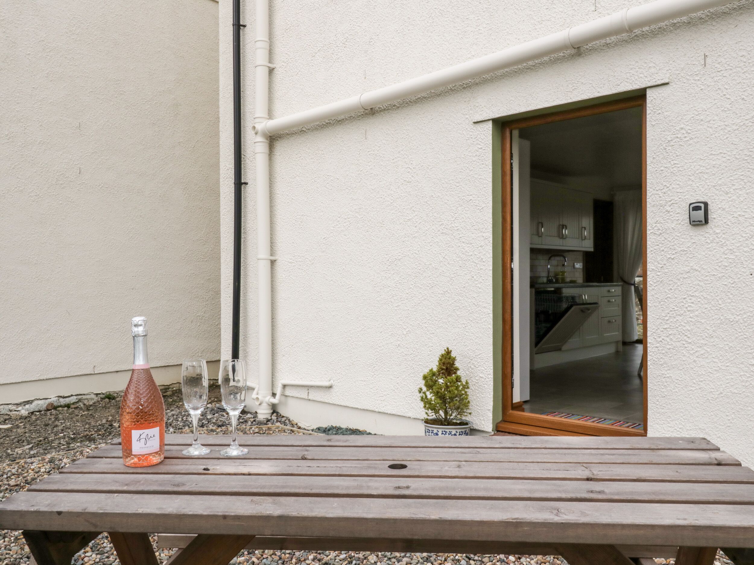 A wooden table with a bottle and glasses, a small potted plant, and a kitchen visible through an open door.
