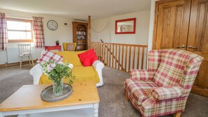 A cozy living room with a yellow sofa, a plaid armchair, a wooden coffee table with a vase of flowers, a bookshelf, and a wooden staircase. - Bramblewood Cottage (Wigton)