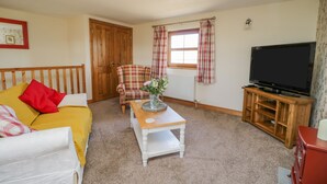 A cozy living room with a yellow sofa, a plaid armchair, a wooden coffee table, and a television on a wooden stand. - Bramblewood Cottage (Wigton)