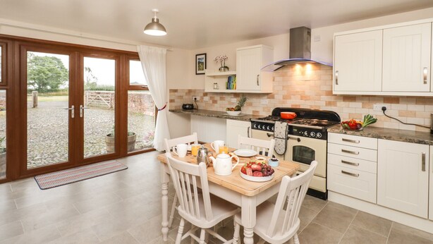 A kitchen with white cabinets, a wooden dining table, and a large window with a view of a garden. - Bramblewood Cottage (Wigton)