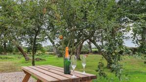 A wooden picnic table with a bottle and two glasses on it, set in a grassy area with trees in the background. - Bramblewood Cottage (Wigton)