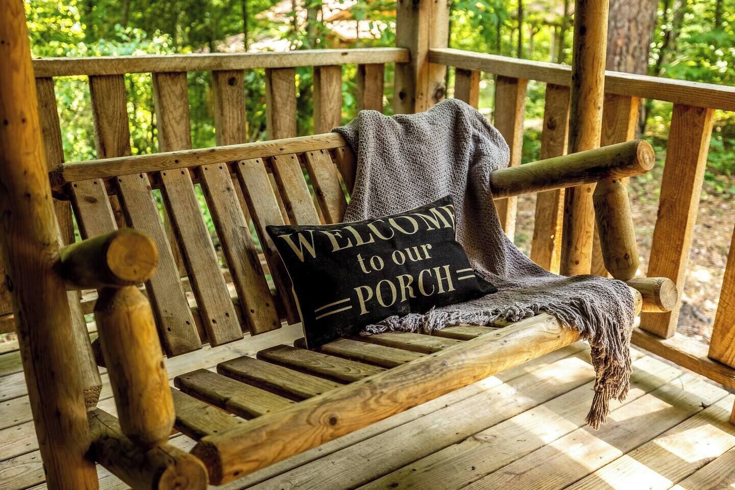 A wooden bench with a 'Welcome to our Porch' sign, set on a wooden deck with a natural backdrop.