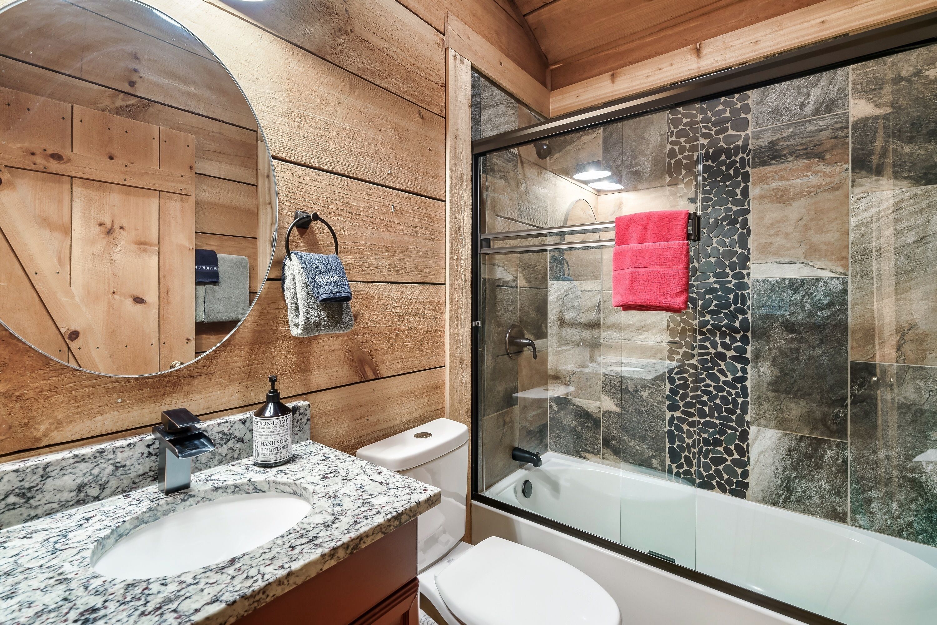 Une salle de bain avec des murs en bois, un lavabo en granit, des toilettes et une douche en verre, avec une serviette rouge.