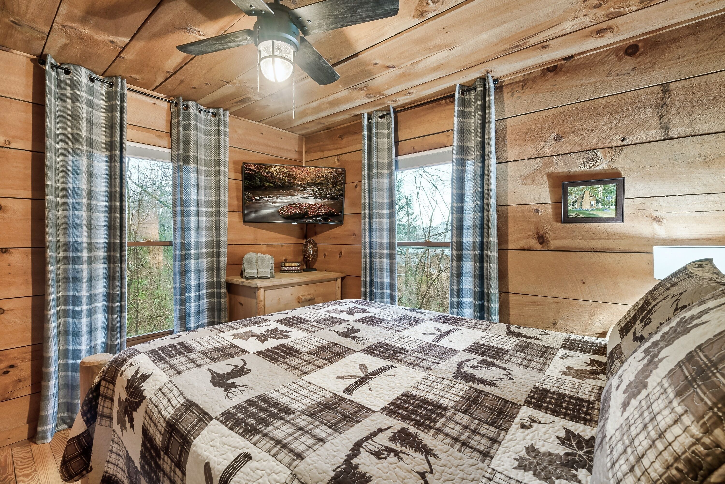 A wooden cabin room with a quilted bedspread, plaid curtains, a ceiling fan, a TV, and a framed picture on the wall.