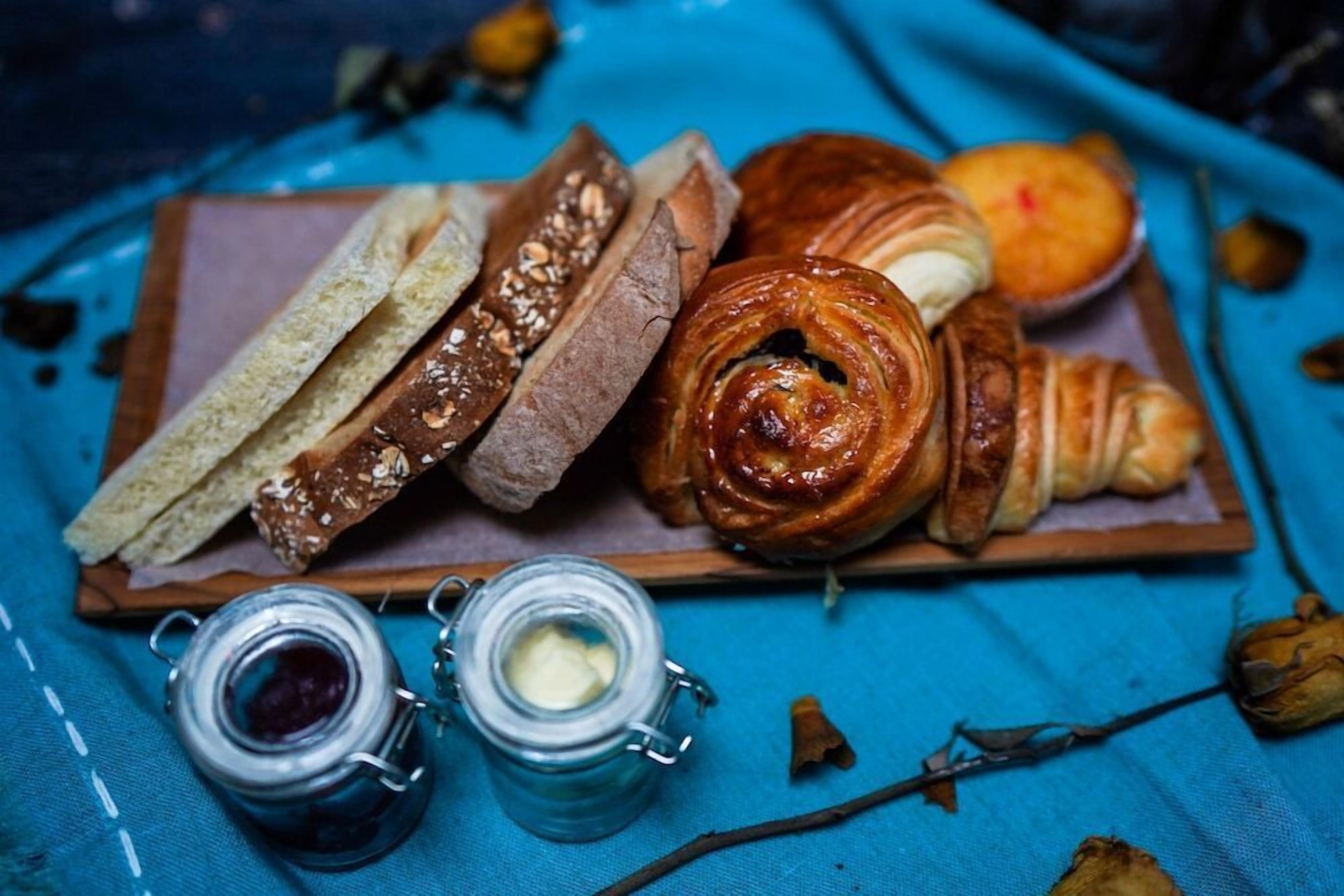 A wooden board with various breads and pastries, two jars of spreads, and a blue cloth background.
