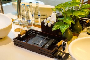 A bathroom vanity with a large white sink, a mirror, and a green plant with white flowers. There are several bottles of water and a small black box with a white label. - Maca Villas Spa Umalas (Kerobokan)