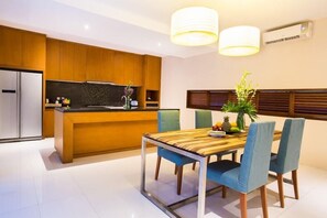 A modern kitchen with wooden cabinets, a stainless steel refrigerator, a built-in oven, and a dining area with a wooden table and blue chairs. - Maca Villas Spa Umalas (Kerobokan)