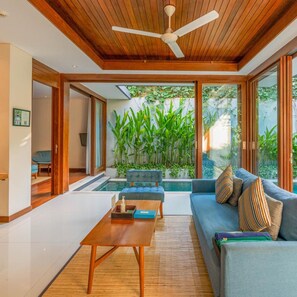 A modern living room with a wooden ceiling, a ceiling fan, a pool, and a view of greenery. - Maca Villas Spa Umalas (Kerobokan)