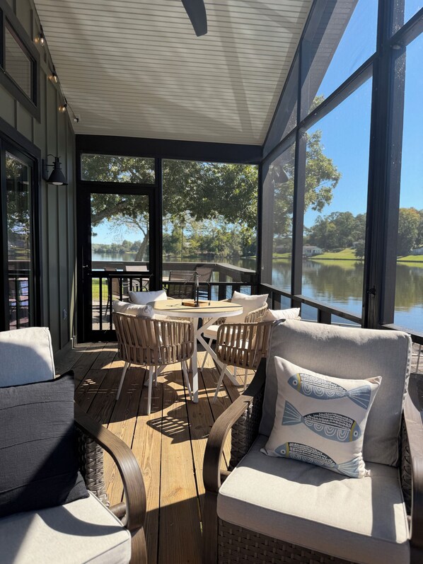 A covered outdoor seating area with a table and chairs, overlooking a body of water and greenery.