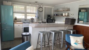 Cocina compacta con refrigerador color turquesa, microondas y estufa. Hay taburetes de bar y una silla azul con cojín.